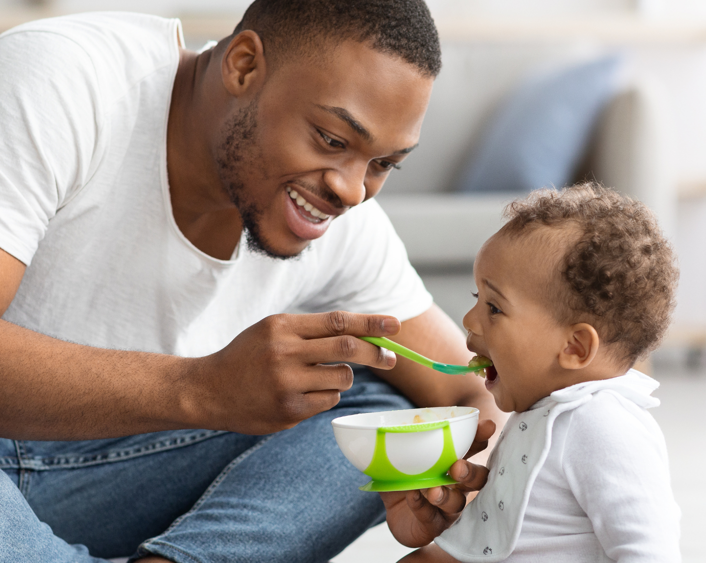 A man is seen smiling, sat cross legged on the floor. He is leaning forward, holding a small plastic bowl in one hand and a spoon in the other. He is offering the spoon of food to a baby who has their mouth open.